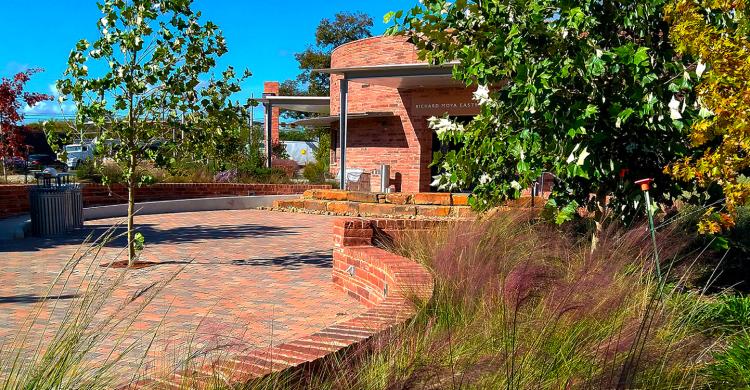 Red brick walkway leads to low circular building, with trees and grasses throughout the area.